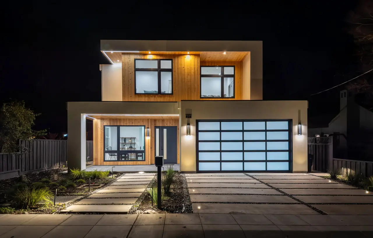 Modern two-story home at night with wood siding and a lit glass garage door
