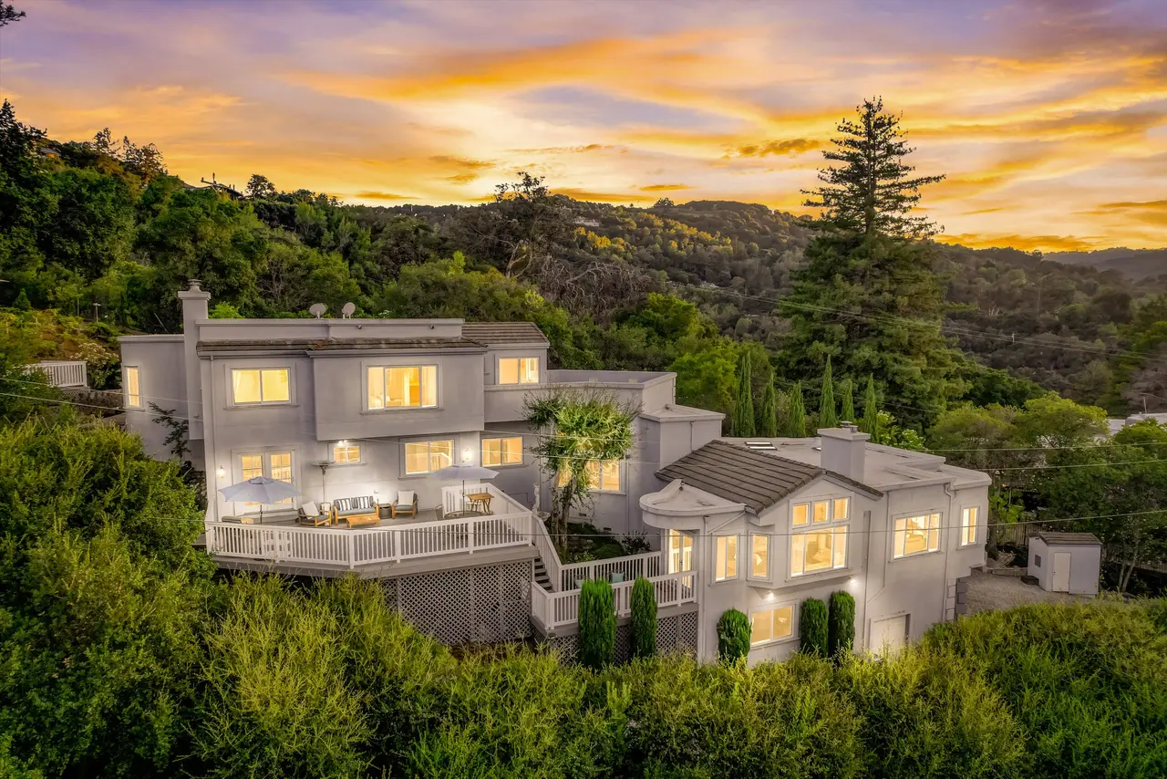 Hillside home at sunset with glowing windows, a large deck, and wooded hills behind